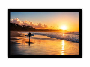 Lone surfer standing on a beach at sunset with golden reflections across calm ocean waves