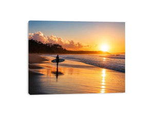 Lone surfer standing on a beach at sunset with golden reflections across calm ocean waves
