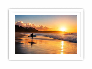 Lone surfer standing on a beach at sunset with golden reflections across calm ocean waves