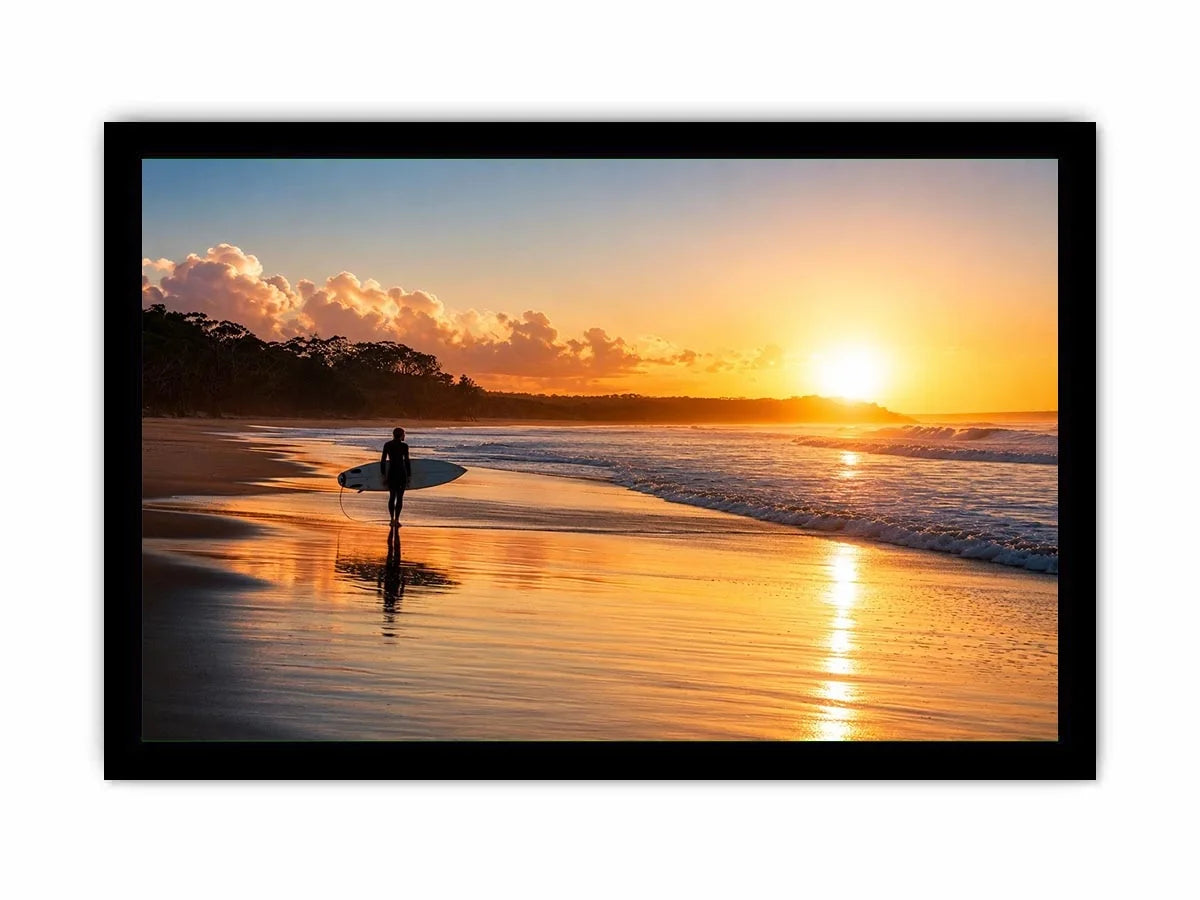 Lone surfer standing on a beach at sunset with golden reflections across calm ocean waves