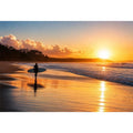 Lone surfer standing on a beach at sunset with golden reflections across calm ocean waves