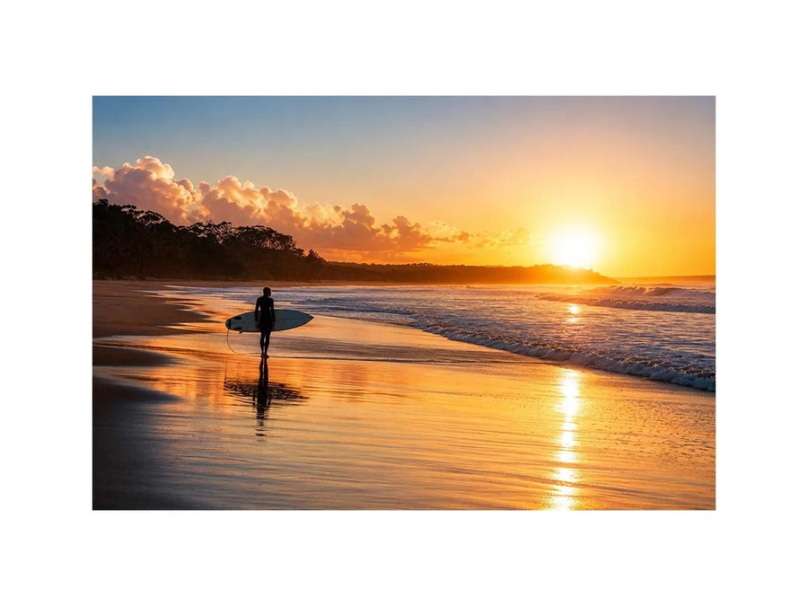 Lone surfer standing on a beach at sunset with golden reflections across calm ocean waves