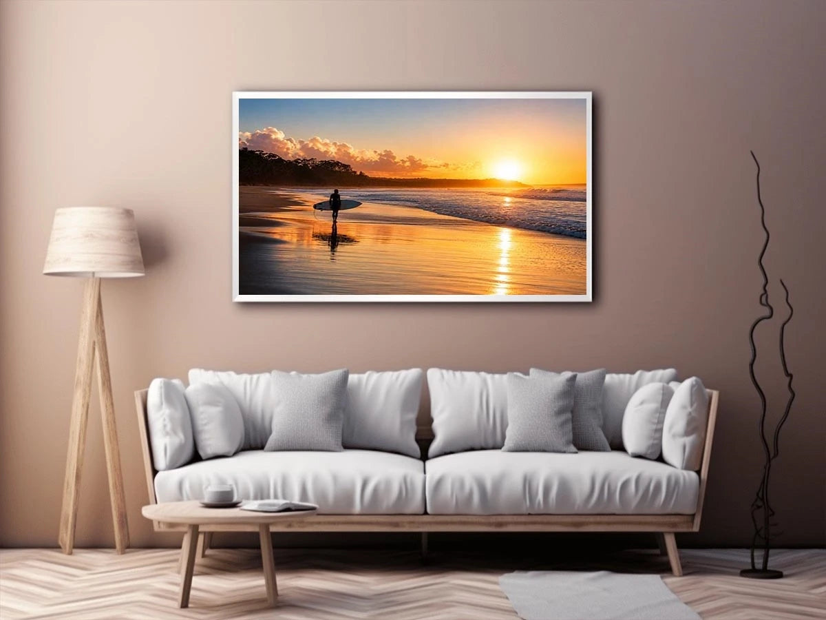 Lone surfer standing on a beach at sunset with golden reflections across calm ocean waves