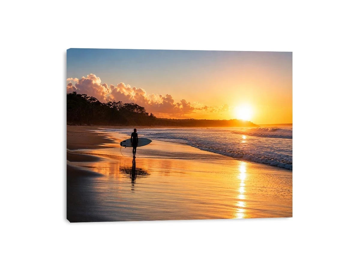 Lone surfer standing on a beach at sunset with golden reflections across calm ocean waves