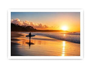 Lone surfer standing on a beach at sunset with golden reflections across calm ocean waves