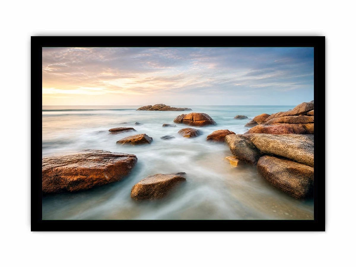 Serene coastal landscape wall art featuring smooth ocean waves, natural shoreline rocks, pastel sunrise sky, and luxury long exposure beach photography for modern coastal interiors
