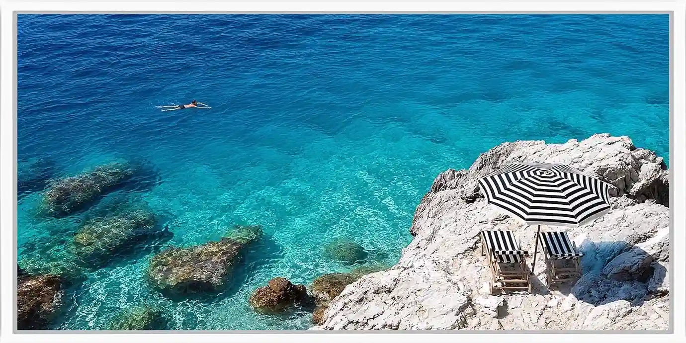 Turquoise coastal cove canvas art with white rocks, striped umbrella and clear sea.